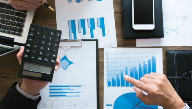 Hands using a calculator and pointing at printed bar and line charts, illustrating investment comparison