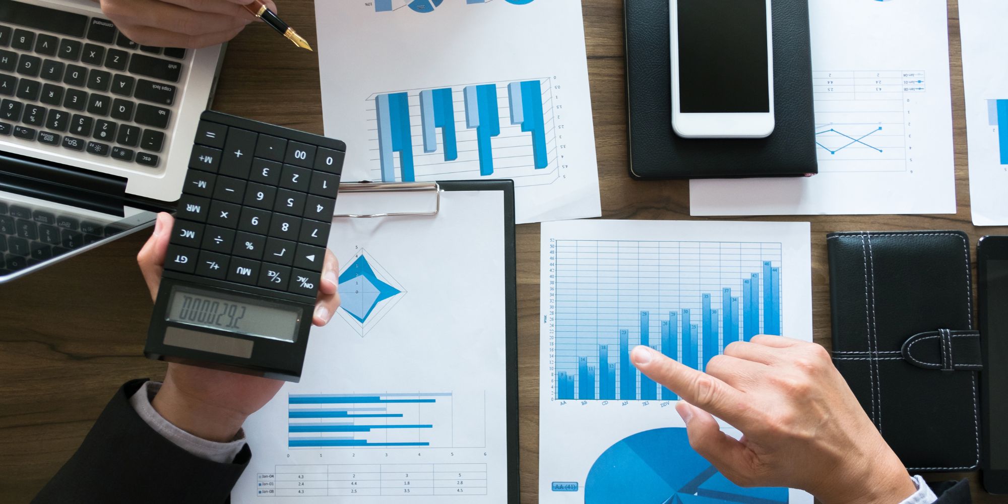 Hands using a calculator and pointing at printed bar and line charts, illustrating investment comparison